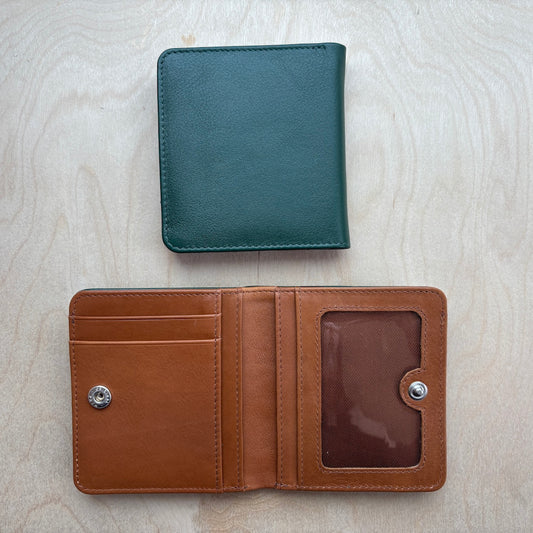 Two leather bifold wallets shown on a light wood background: one closed in hunter green at the top and one open in tan brown at the bottom, displaying card slots and an ID window.