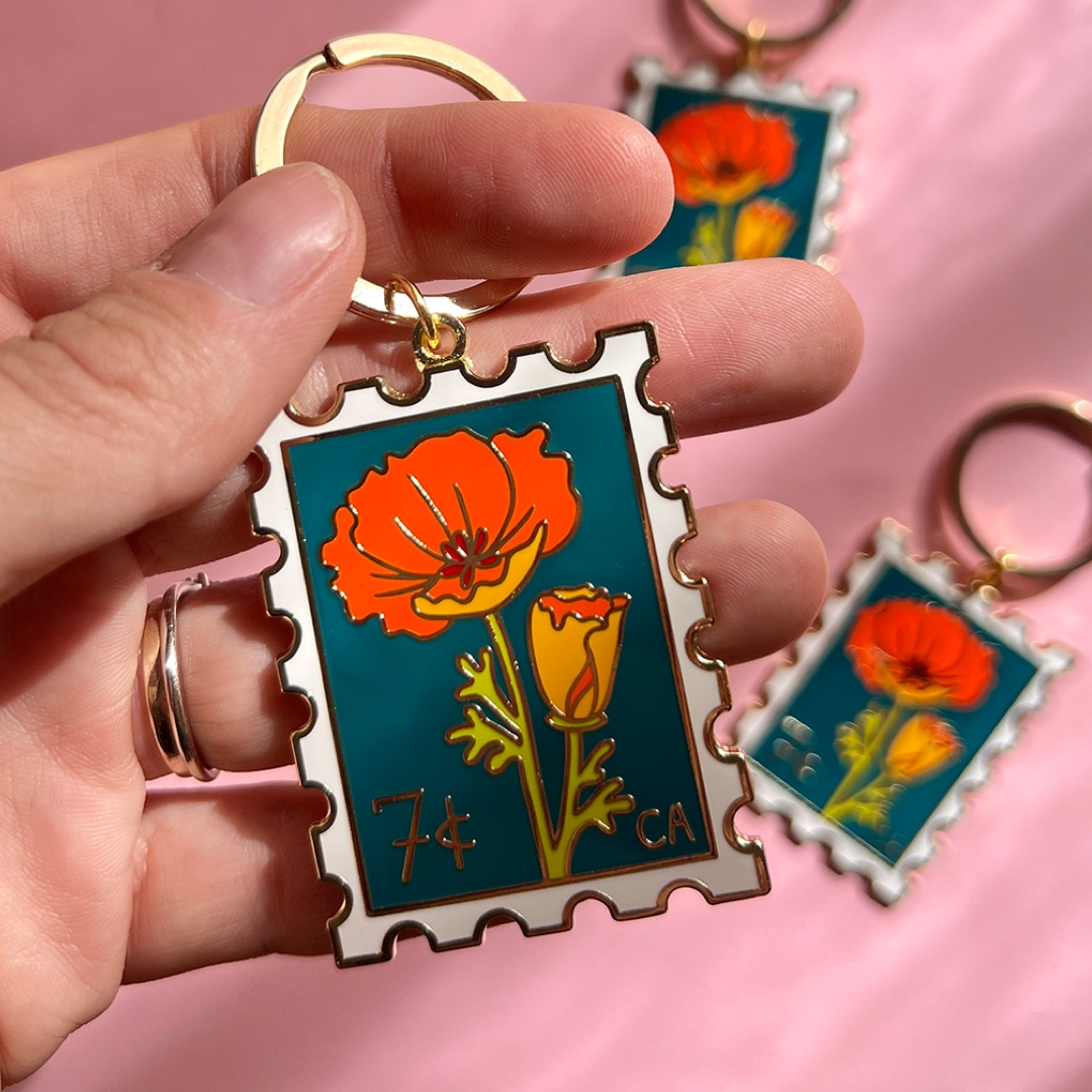 Floral keychain with '7 cent CA' poppy stamp design held by a hand against a pink background