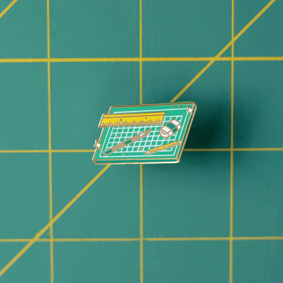 Pin with the design of a mini cutting mat, washi tape, eraser, x-acto knife, and pencil