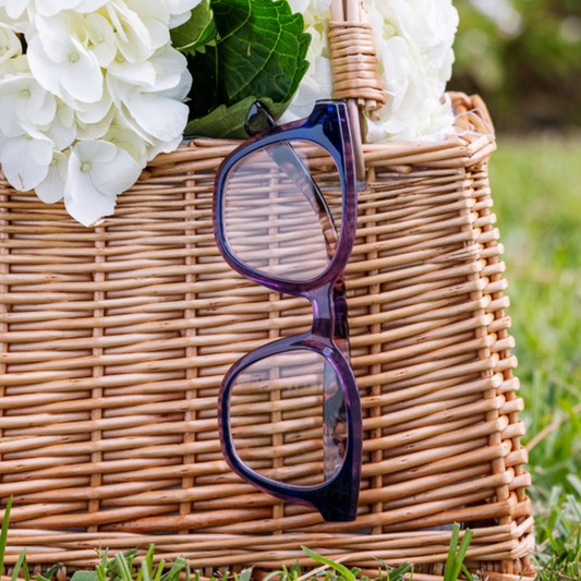Purple-framed glasses on a wicker basket with white flowers and green leaves outdoors.