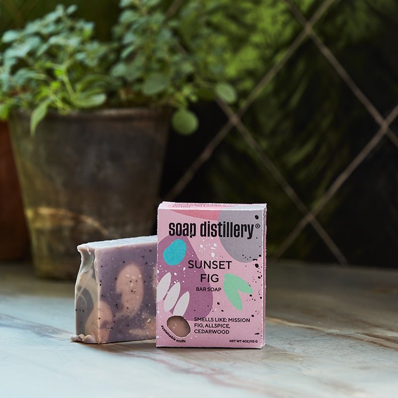 Bar of soap and its packaging labeled 'Soap Distillery Sunset Fig' on a surface with a plant in the background.