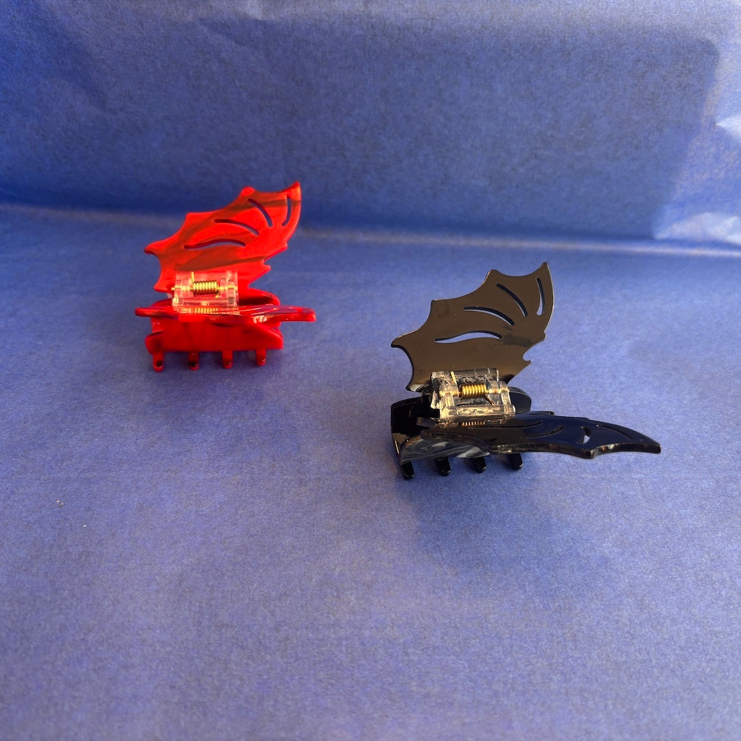 Two decorative bat shaped claw hair clips, one red and one glossy black, displayed on a textured blue background.
