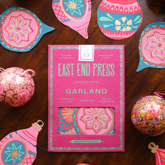 A pink East End Press Garland printed paper sheet with a window showing colorful patterned paper inside, styled on a dark wood surface with decorative paper ornaments around it.