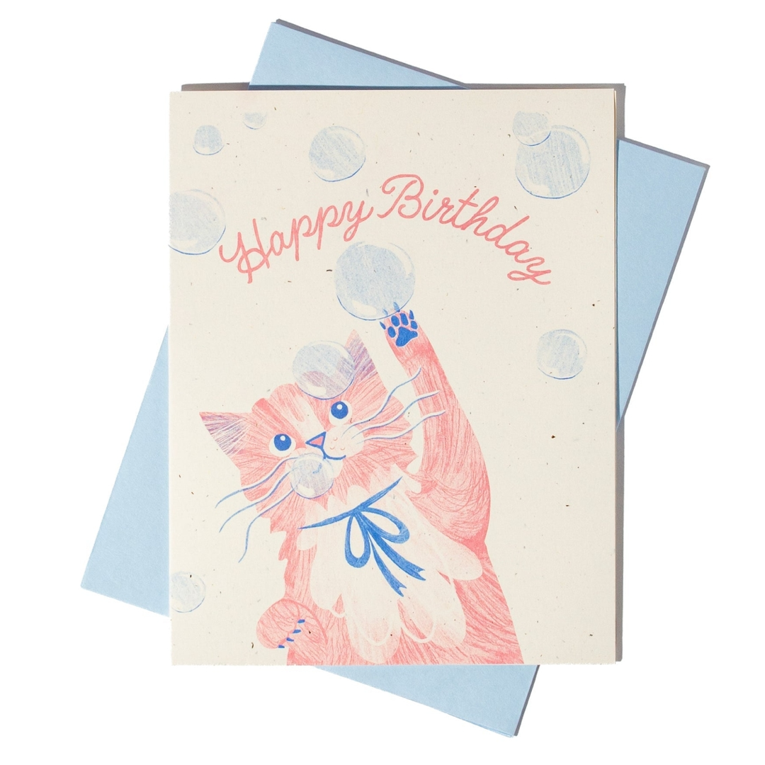 A square birthday card with a cream background, featuring a pink and white cat reaching up to bubbles with its paw and the words 'Happy Birthday' in pink script. The card is layered over a light blue envelope on a white background.