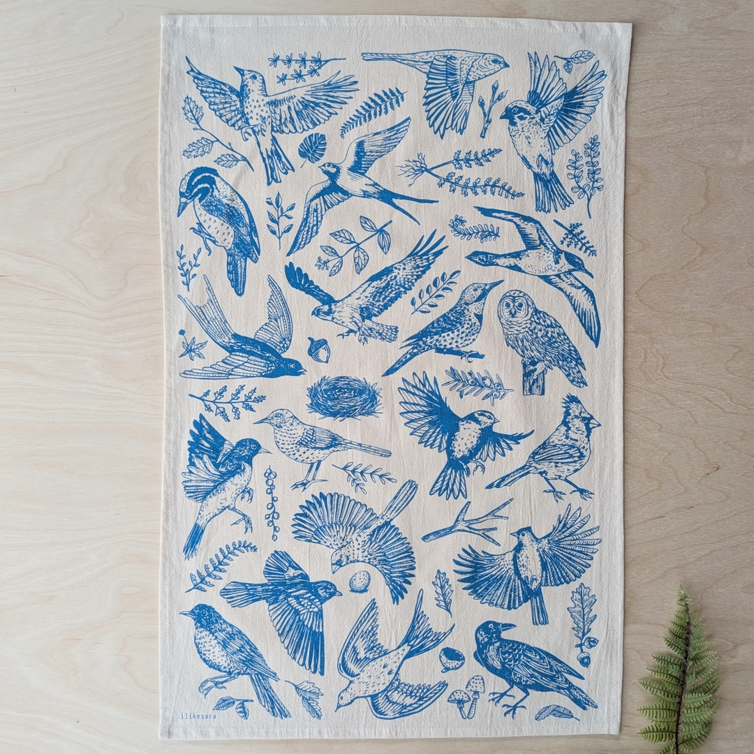A square white fabric tea towel featuring a blue bird and nature print, laid flat on a light wood background with a small fern leaf at the corner.