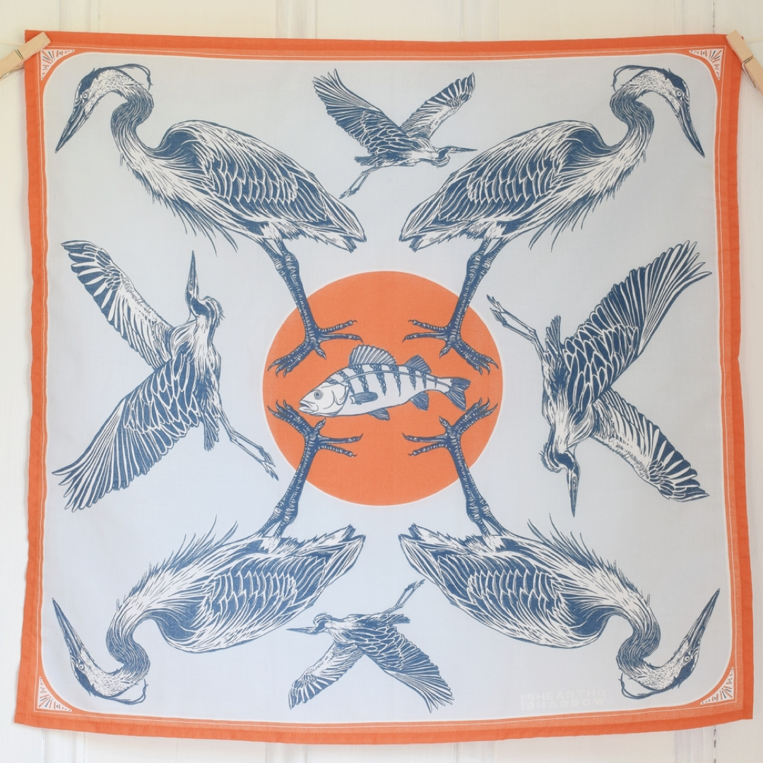 A square scarf with an orange border, featuring a central orange circle with a striped fish and surrounded by eight blue heron illustrations, hanging against a white wood-paneled wall.