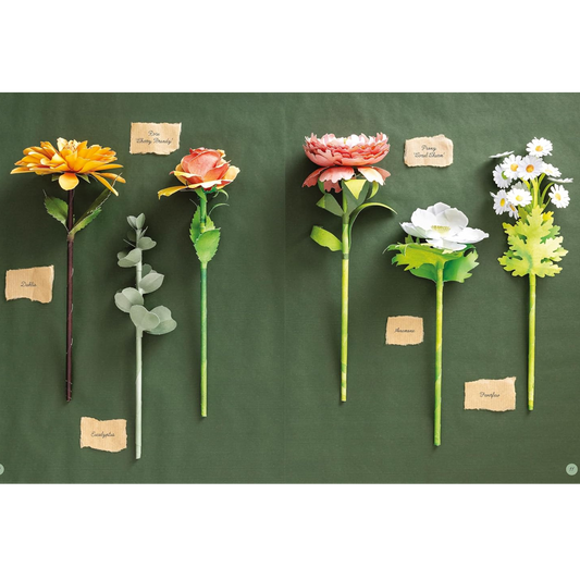 Six artificial paper flowers with green stems are arranged on a dark green chalkboard background, each with a small handwritten label card. The flowers include yellow, peach, coral, white, and daisy varieties.