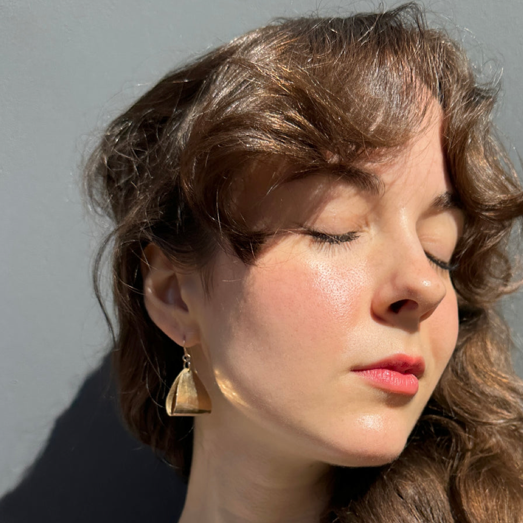 A close-up portrait of a woman with closed eyes wearing a small gold geometric dangle earring, set against a plain gray background with dramatic side lighting.