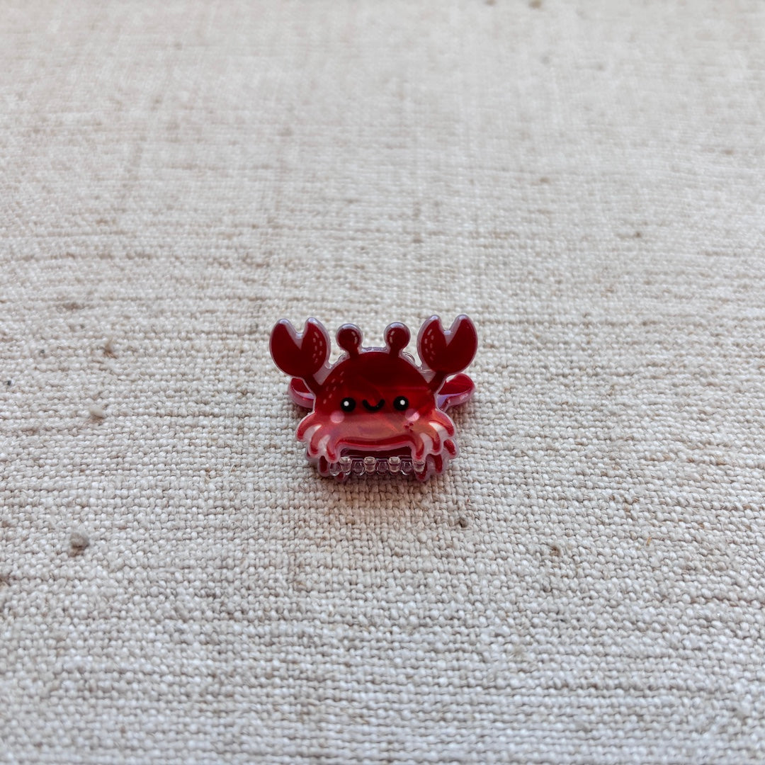A small red crab-shaped hair clip displayed on a textured beige fabric background.