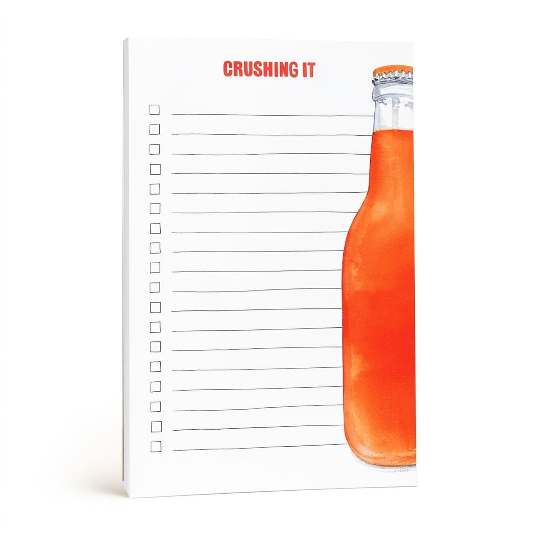 A white lined notepad with the red text 'CRUSHING IT' at the top and a large orange soda bottle illustration on the right side, shown on a clean white background.