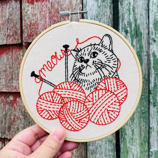 Embroidery hoop with a cat and yarn design held against a wooden background