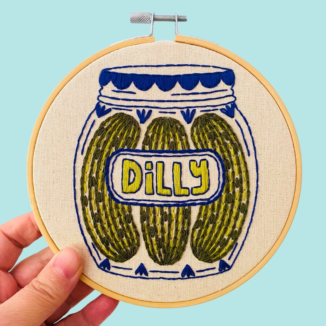 A hand holds a wooden embroidery hoop featuring a stitched design of three pickles in a jar with the word 'DILLY' on the label, set against a light blue background.