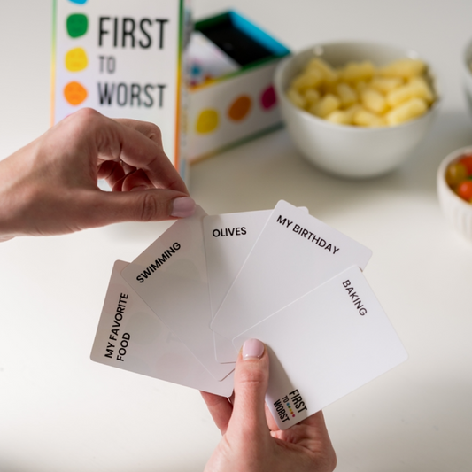 Hands holding playing cards with text, 'First to Worst' game box in the background