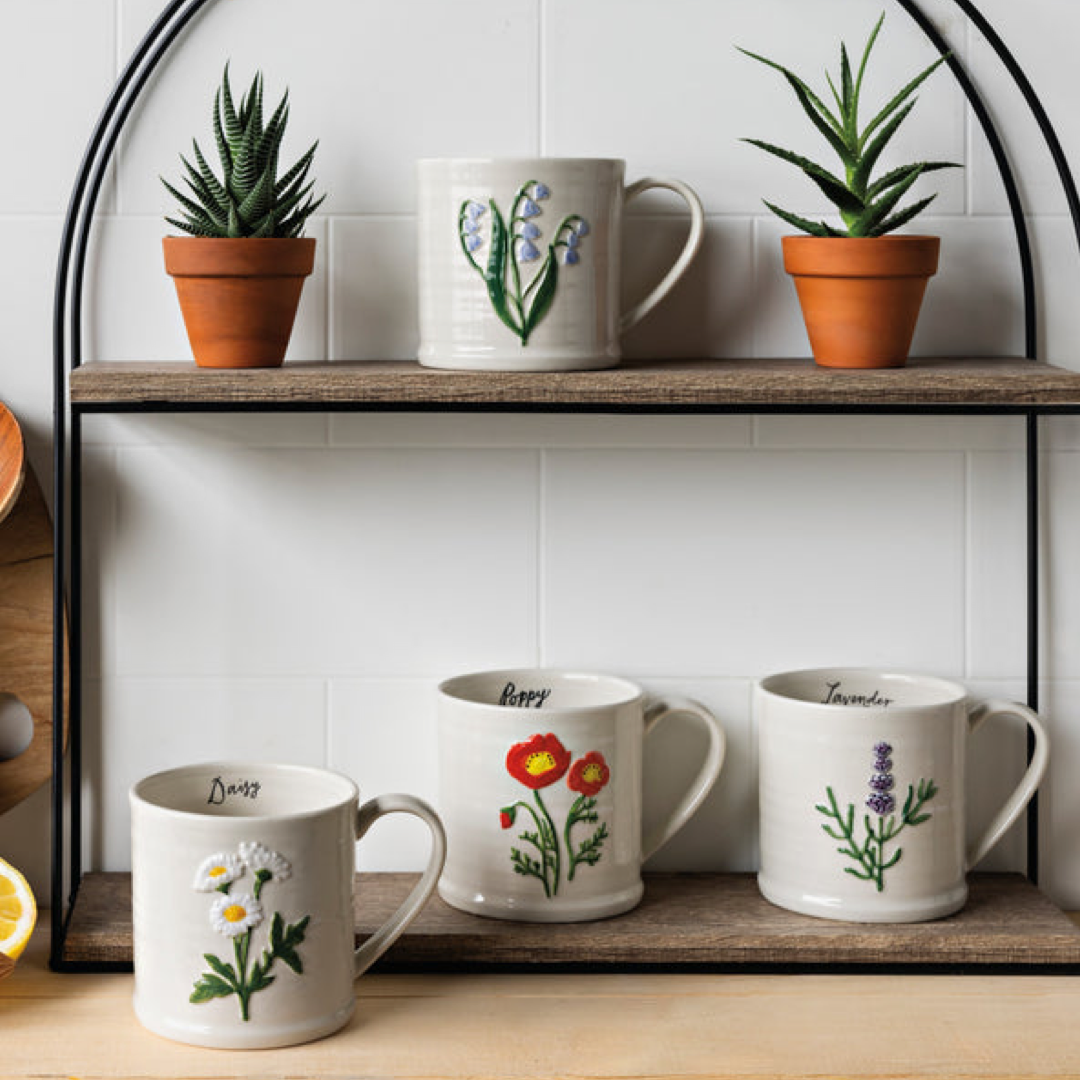 Four white ceramic mugs with different colorful botanical illustrations are arranged on two rustic wooden shelves against a white tiled wall, with potted succulents on top.