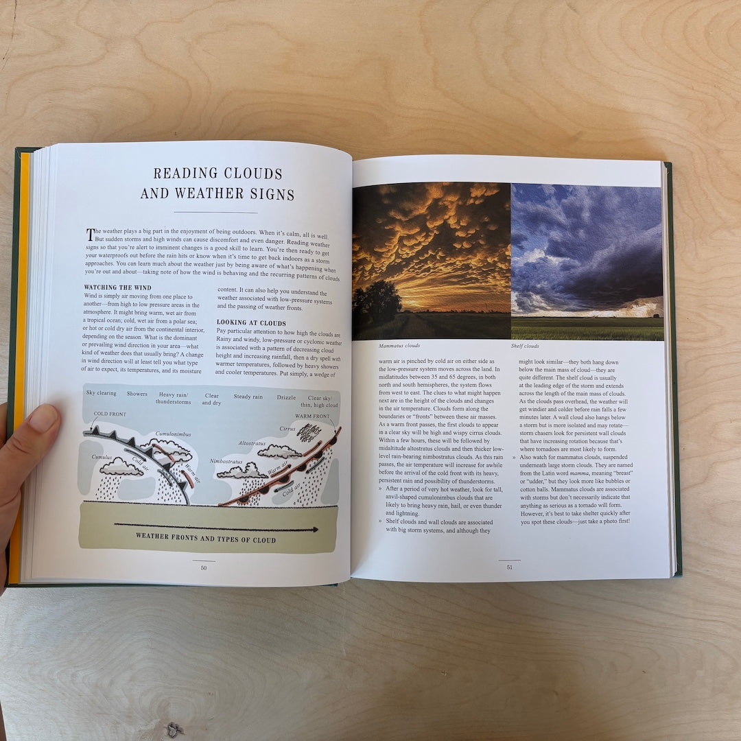 An open book showing a weather-related article titled 'Reading Clouds and Weather Signs,' with text, diagrams, and two landscape photos on the pages. A hand is holding the left page, and the book is placed on a light wood surface.