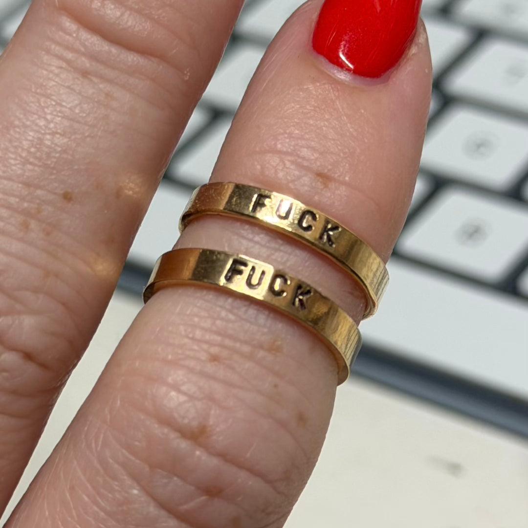 Two gold band rings engraved with the word 'FUCK' stacked on a person's ring finger, shown against a blurred keyboard background.