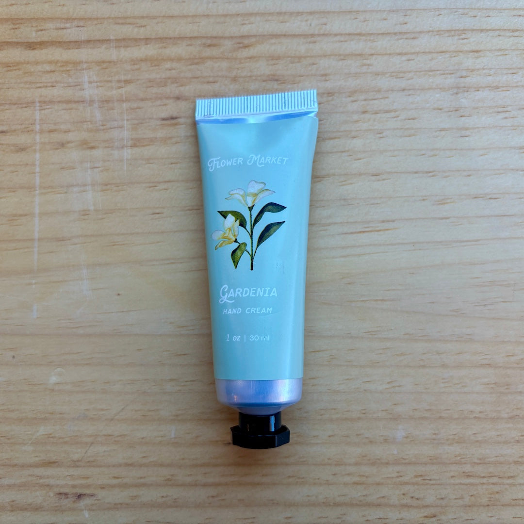 A light green squeeze tube of Flower Market Gardenia Hand Cream with a black cap, placed on a light wood grain surface.