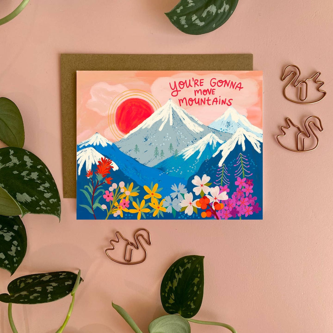 Card with mountain and floral design and text that says you're gonna move mountains on it on a pink background with decorative elements.