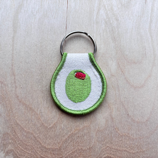 An embroidered stuffed olive keychain featuring a white fabric body with green stitched edges, a green olive center, and a small red pepper detail, attached to a silver metal split ring and placed on a light wood background.
