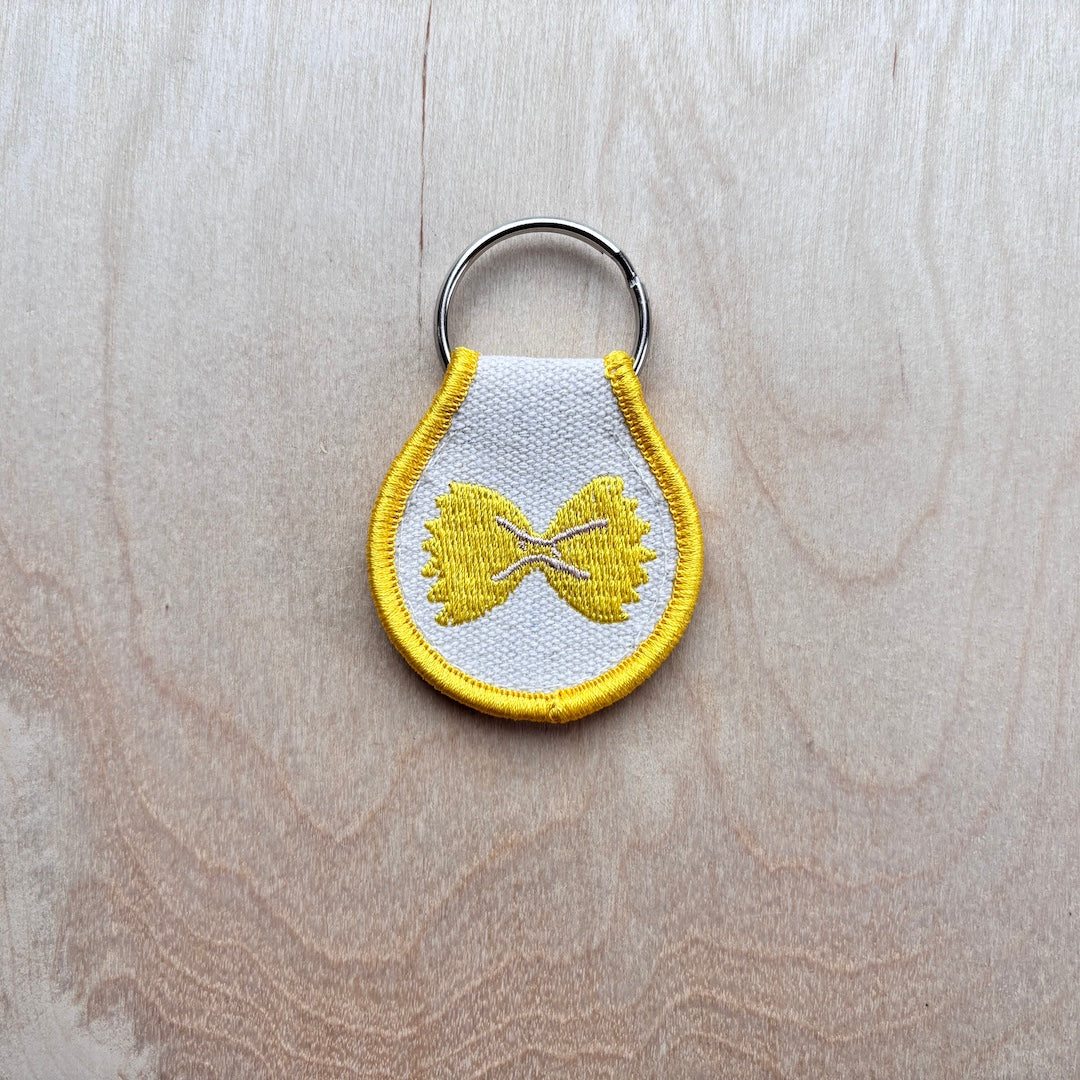 A small embroidered keychain with a white fabric body, yellow border, and yellow bowtie pasta motif, attached to a silver metal key ring, placed on a light wood surface.