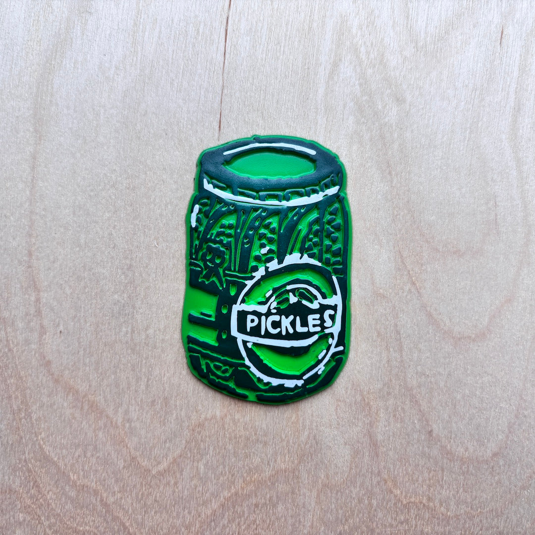 A green fridge magnet shaped like a pickle jar with a white circular logo reading 'PICKLES', placed on a light wood grain background.