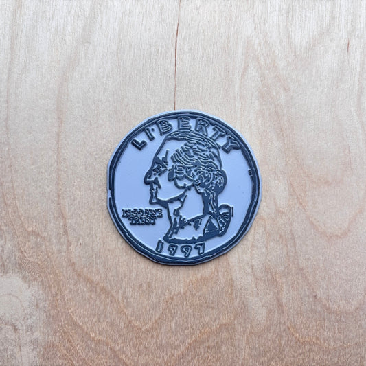 A circular fridge magnet designed to look like a US quarter coin, featuring a dark blue-gray profile of George Washington with 'LIBERTY' and '1997' embossed on a light blue-gray background, placed on a light wood grain surface.