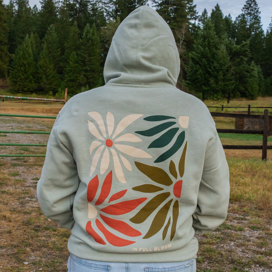 A person standing outdoors in a wooded field, viewed from behind, wearing a sage hoodie with a large retro floral graphic on the back in cream, red, olive green, and dark green. The text 'IN FULL BLOOM' is printed near the lower back.