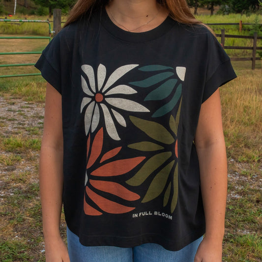 A person wearing a black graphic t-shirt with a retro floral design in white, orange, and green, standing outdoors in a grassy field.