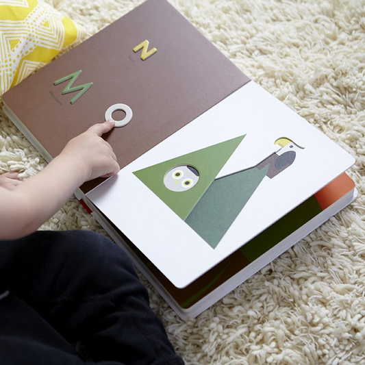 Child's hand interacting with a book featuring letters and shapes on a carpeted floor.