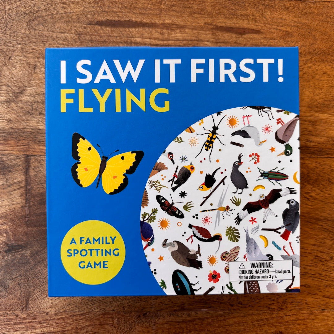 The board game box for 'I Saw It First! Flying' is shown flat on a wooden surface. The box features a bright blue background with yellow and white text, a yellow and black butterfly, and a circular white pattern showing various flying insects and birds.