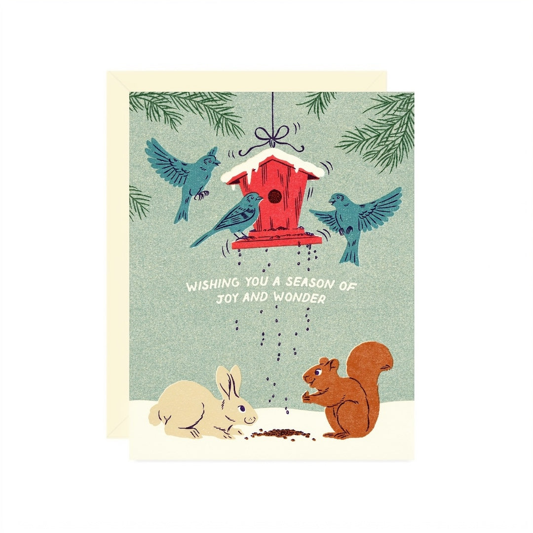 A square greeting card with a cream envelope, featuring a winter garden scene with blue birds around a red birdhouse, a white rabbit, and a brown squirrel. The card reads, 'WISHING YOU A SEASON OF JOY AND WONDER'.