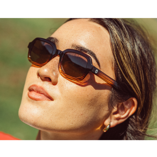A close-up lifestyle shot of a woman wearing dark brown square sunglasses with amber-tinted lenses and tortoiseshell-patterned temples, set against a blurred green background.