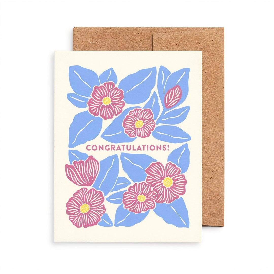 A congratulations greeting card with a cream background, featuring a blue and pink floral illustration with the word 'CONGRATULATIONS!' in pink, shown with a brown kraft envelope behind it on a white background.