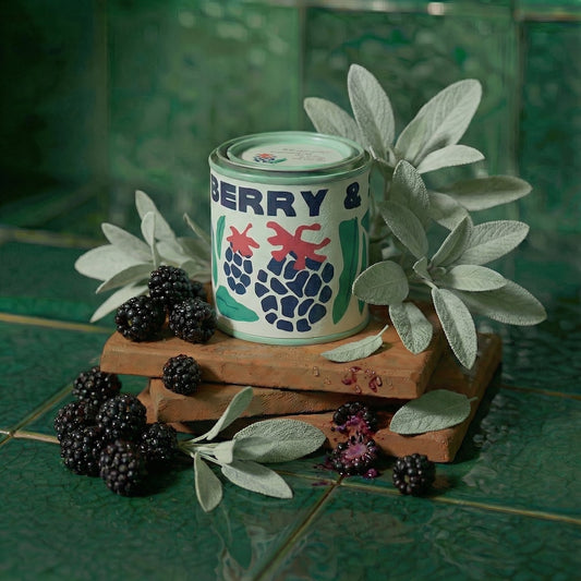 Candle in a tin vessel labeled 'Black Berry & Sage' on a wooden stand surrounded by blackberries and leaves on a green tiled background