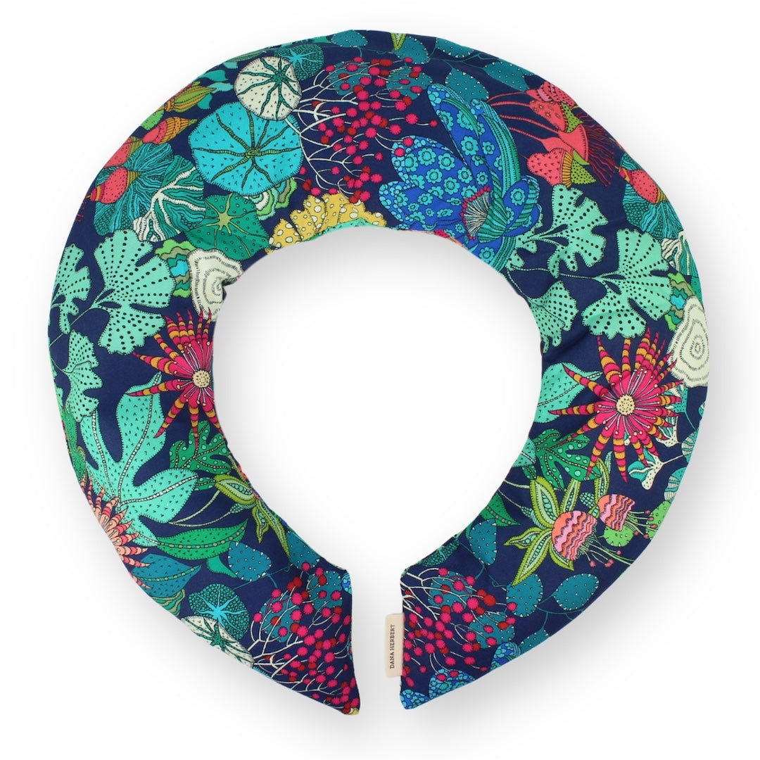 A curved neck pillow featuring a vibrant, multicolored botanical pattern on a dark navy blue background, shown against a plain white background.