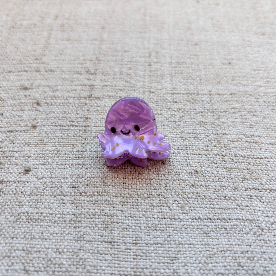A small purple cartoon octopus hair clip with a smiling face and tiny yellow dots, resting on a textured beige linen fabric background.