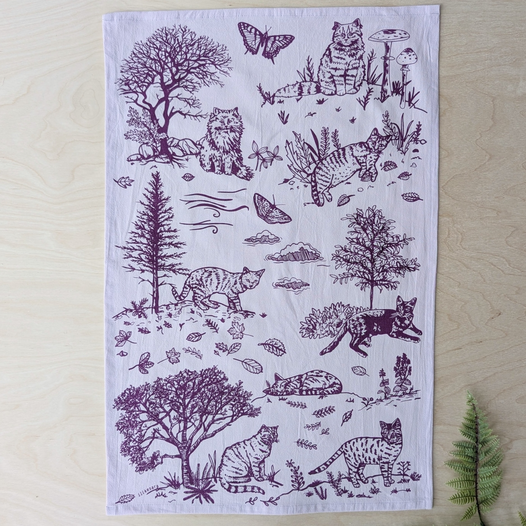 A rectangular white fabric tea towel with a dark purple printed design of various cats, trees, and butterflies, laid flat on a light wood background.