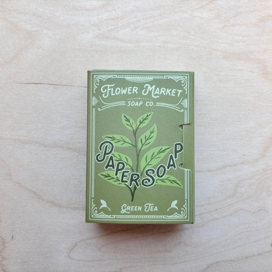 A rectangular box of Flower Market Soap Co. Paper Soap in Green Tea scent, featuring a green and white illustrated design, placed on a light wood grain surface.