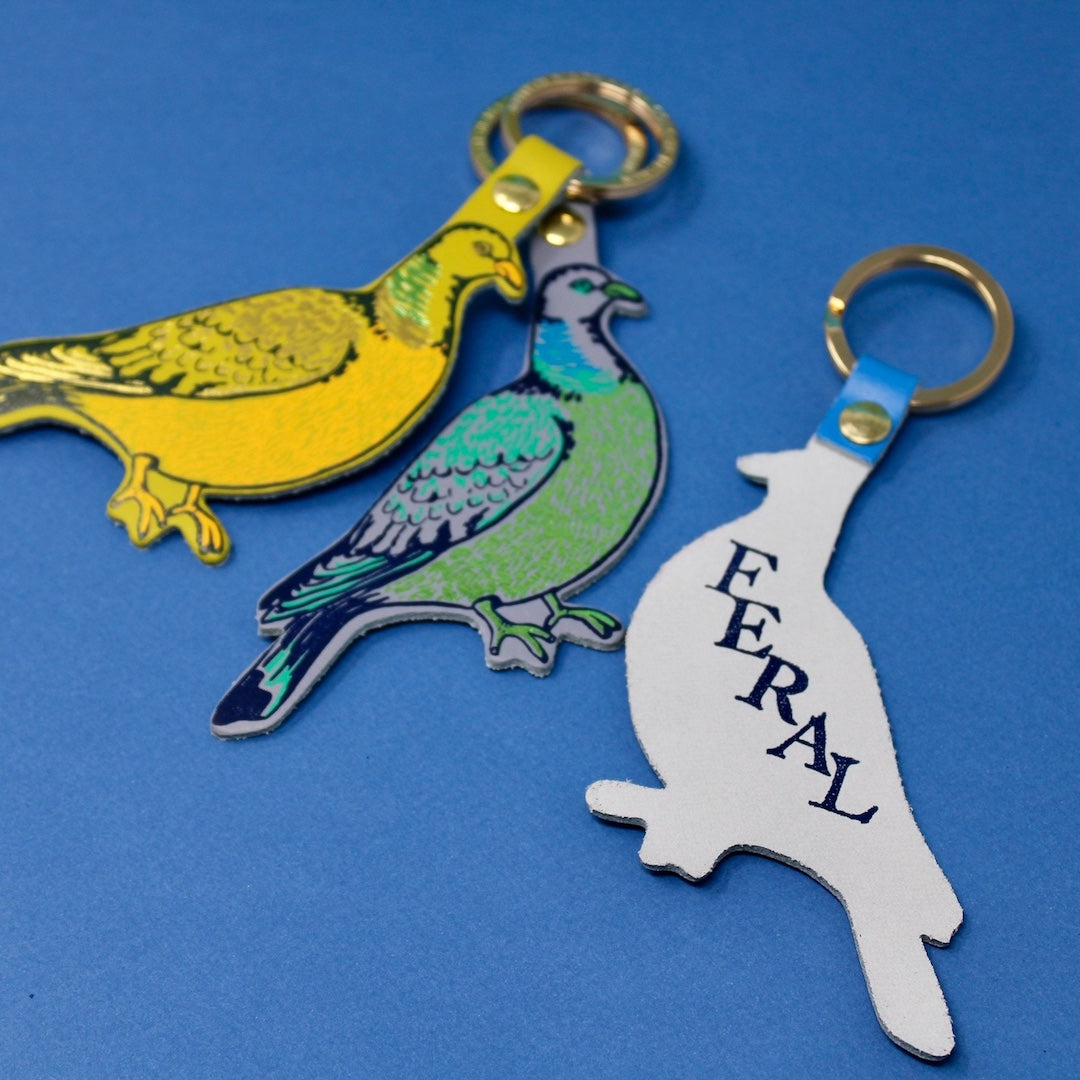 Three bird-shaped keychains on a blue background. The one on the right shows the back side of the keyring which has 'feral' written on it