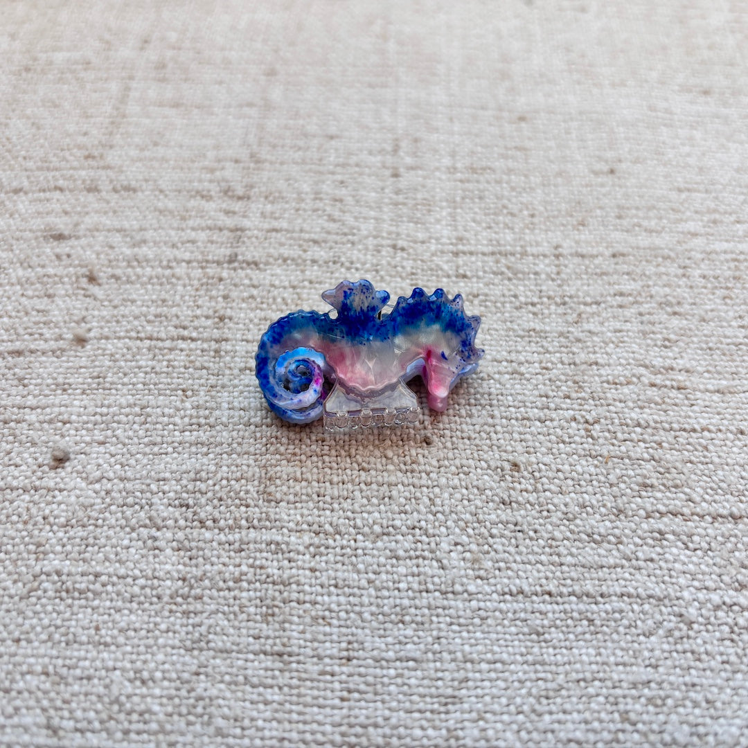 A small seahorse-shaped hair clip with blue, pink, and white details, resting on a textured beige linen fabric background.
