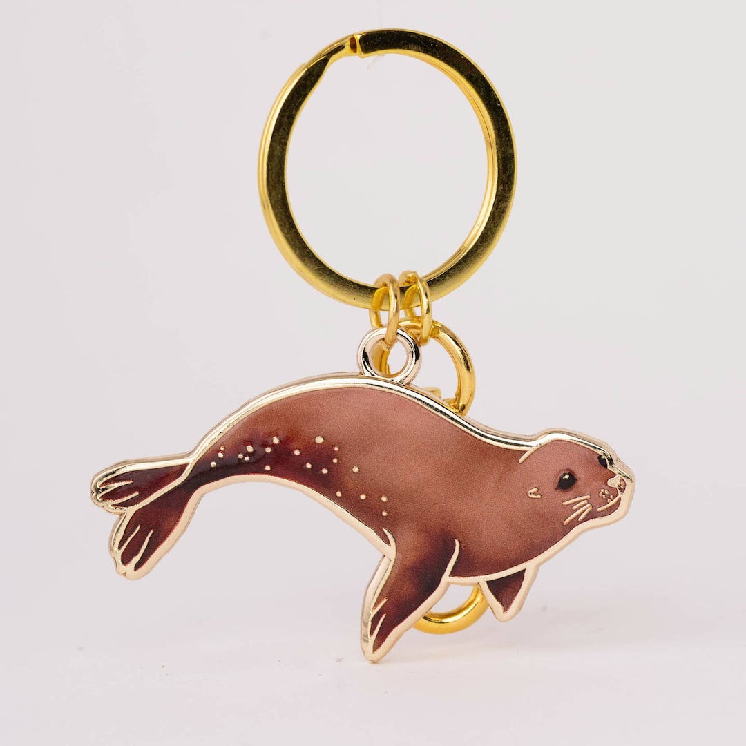 A gold-tone metal keychain featuring an enamel sea lion charm with brown and gold detailing, set against a plain light gray background.