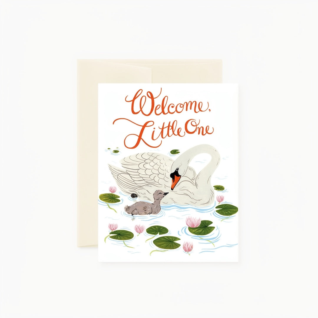 A square greeting card with a cream envelope behind it, featuring an illustration of a white swan and a gray cygnlet among green lily pads and pink flowers, with the words 'Welcome, Little One' in orange script.