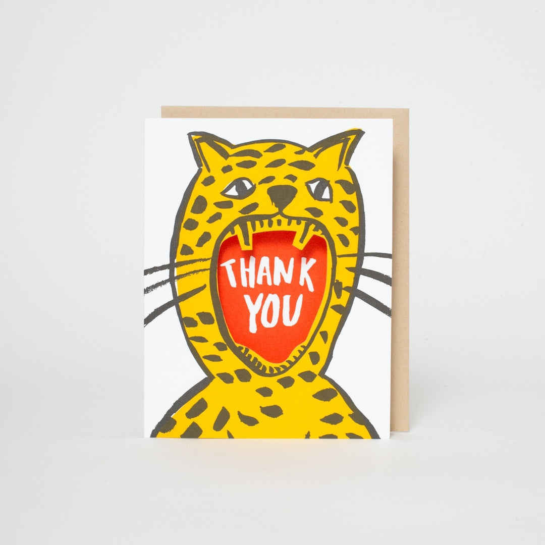 A greeting card featuring a cartoon cheetah with its mouth wide open, revealing the words 'THANK YOU' in white on a red background. The card is shown standing upright next to a light brown envelope against a clean white background.