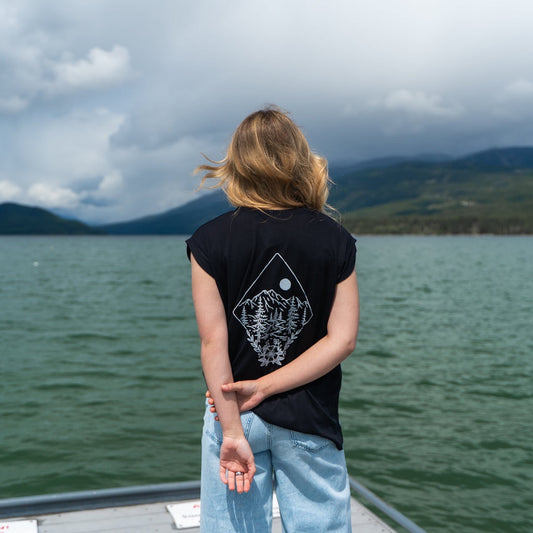 Person by a lake wearing a black t-shirt with a nature design featuring trees, mountains, and the moon