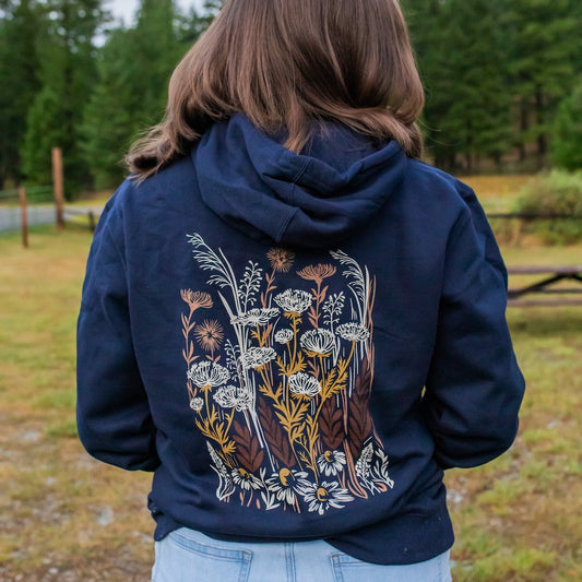 A person viewed from behind wearing a navy blue hoodie with a large back graphic of wildflowers in white, yellow, and brown, set against an outdoor background of trees and grass.