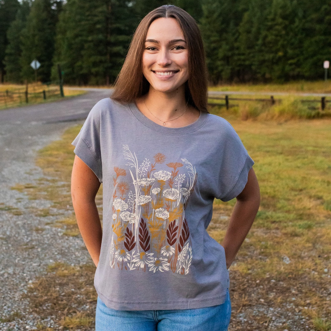 A smiling young woman stands outdoors on a gravel path wearing a heather gray t-shirt with a detailed botanical graphic in white, brown, and gold, paired with blue jeans.