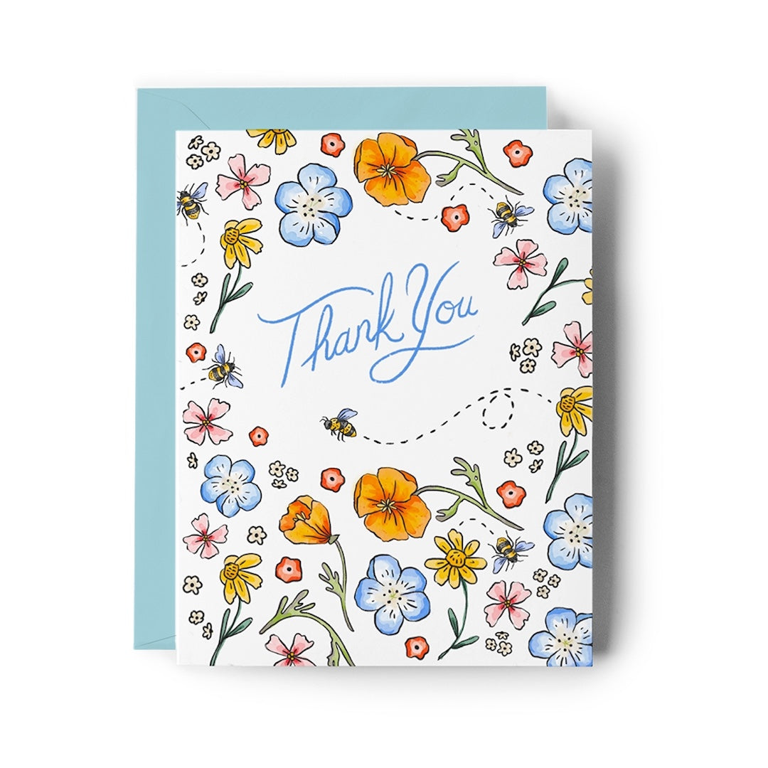 A square white greeting card with a colorful floral border and the words 'Thank You' in blue script, shown with a light blue envelope behind it on a white background.