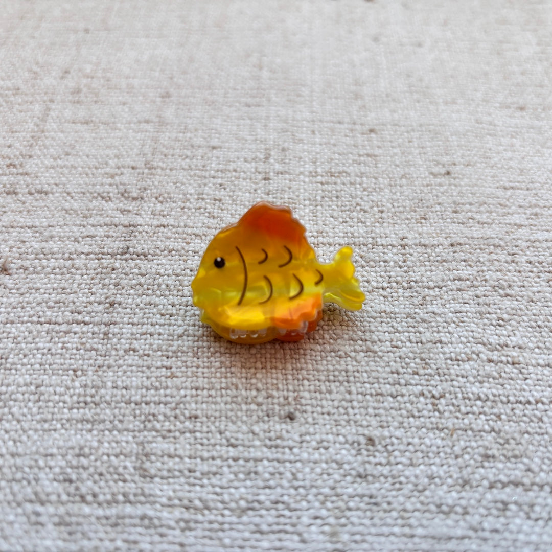 A small yellow and orange fish-shaped hair clip with black scale details, placed on a textured beige fabric background.