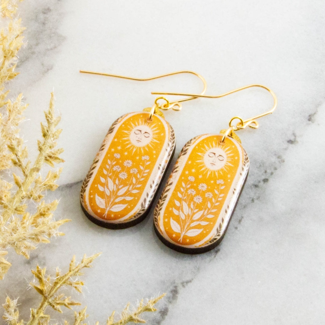 Yellow floral and sun design earrings on a marble surface with decorative plants.