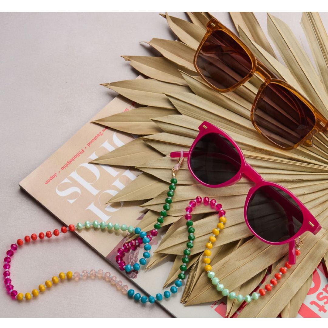Bright pink sunglasses with dark lenses and a colorful beaded strap, styled on a book with dried palm leaves and another pair of sunglasses on a light gray background.